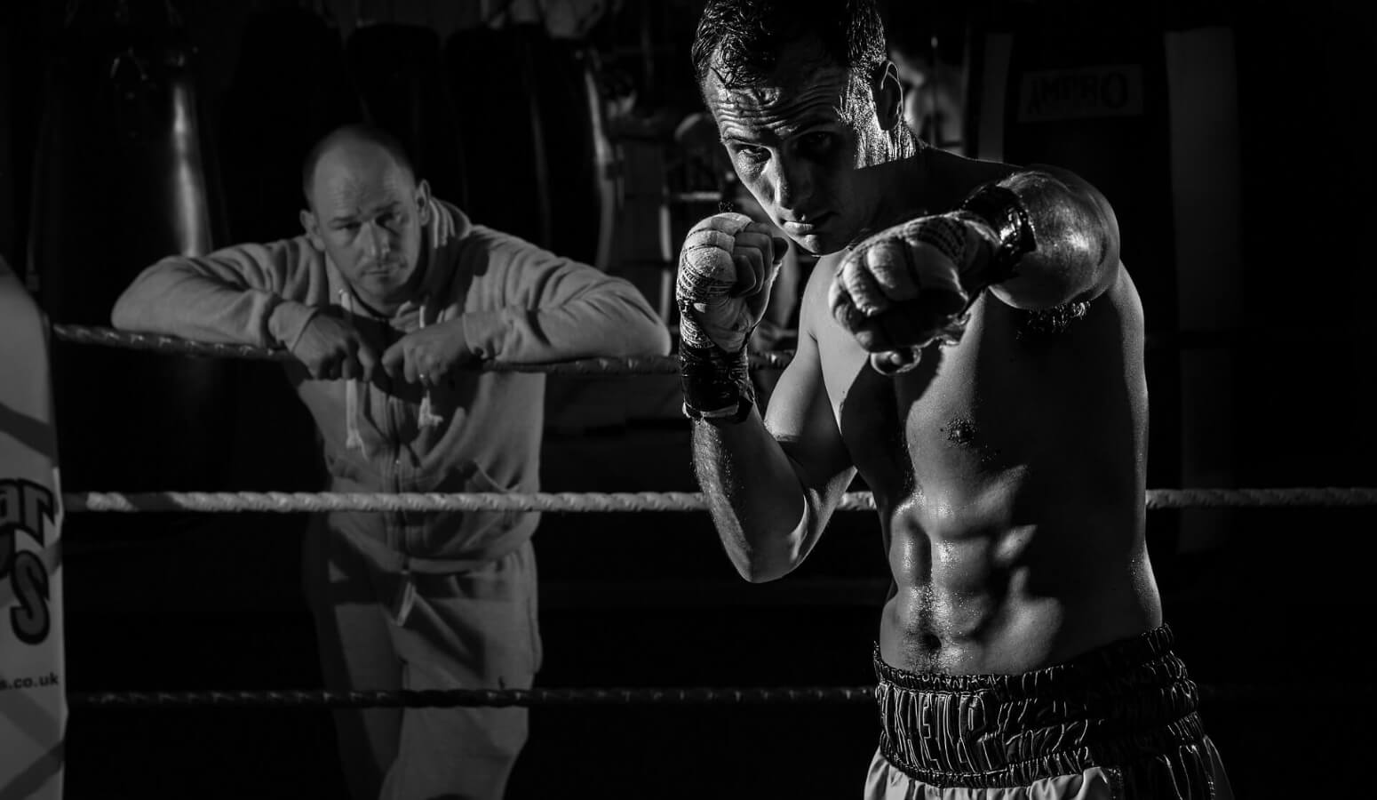 Fighter is standing in the ring and pointing his punch in to a camera while is trainer standing behind him